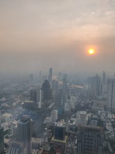 A cityscape view featuring numerous high-rise buildings, with a river running through the middle of the city of Bangkok. The sky is hazy with visible smog, and the sun is setting, casting an orange glow through the haze.