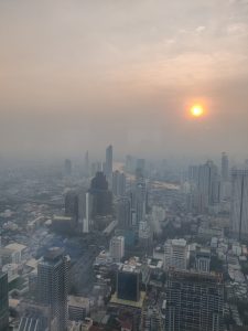 View larger photo: A cityscape view featuring numerous high-rise buildings, with a river running through the middle of the city of Bangkok. The sky is hazy with visible smog, and the sun is setting, casting an orange glow through the haze.