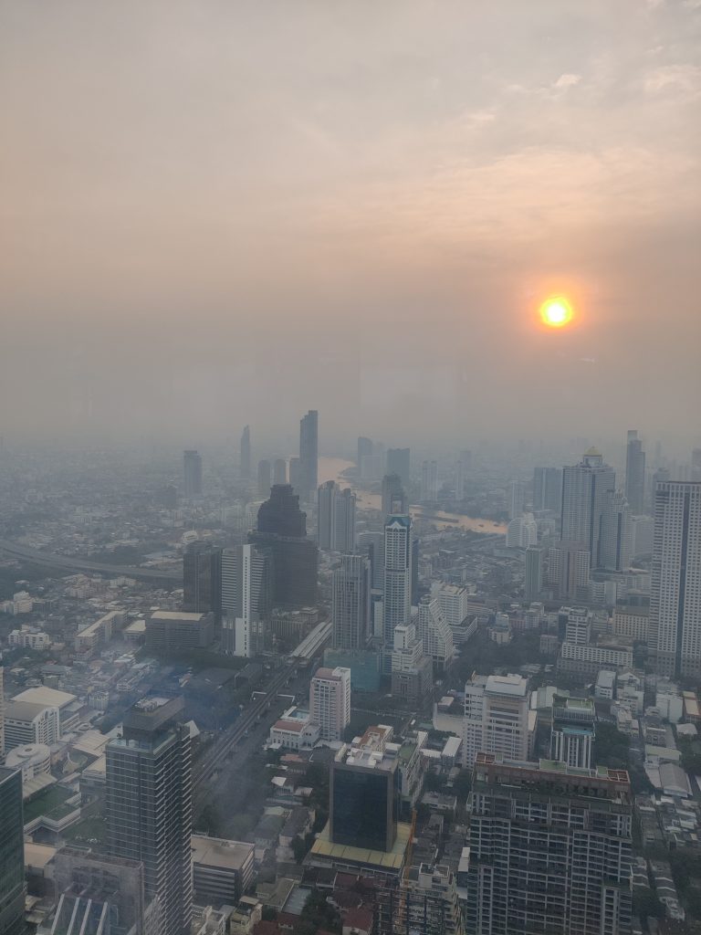 A cityscape view featuring numerous high-rise buildings, with a river running through the middle of the city of Bangkok. The sky is hazy with visible smog, and the sun is setting, casting an orange glow through the haze.