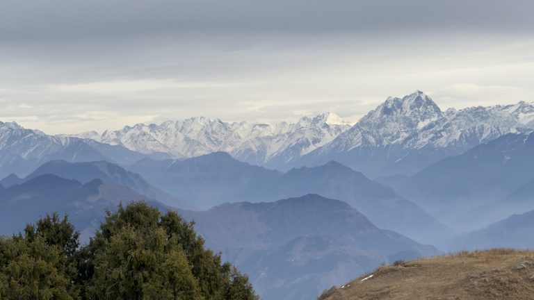 Snow-capped mountains in the distance under a cloudy sky, with rolling hills and green trees in the foreground.