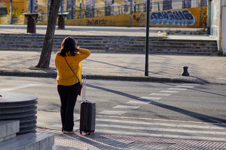 A woman standing with a suitcase on a sidewalk, looking at a city street with pedestrian crossings