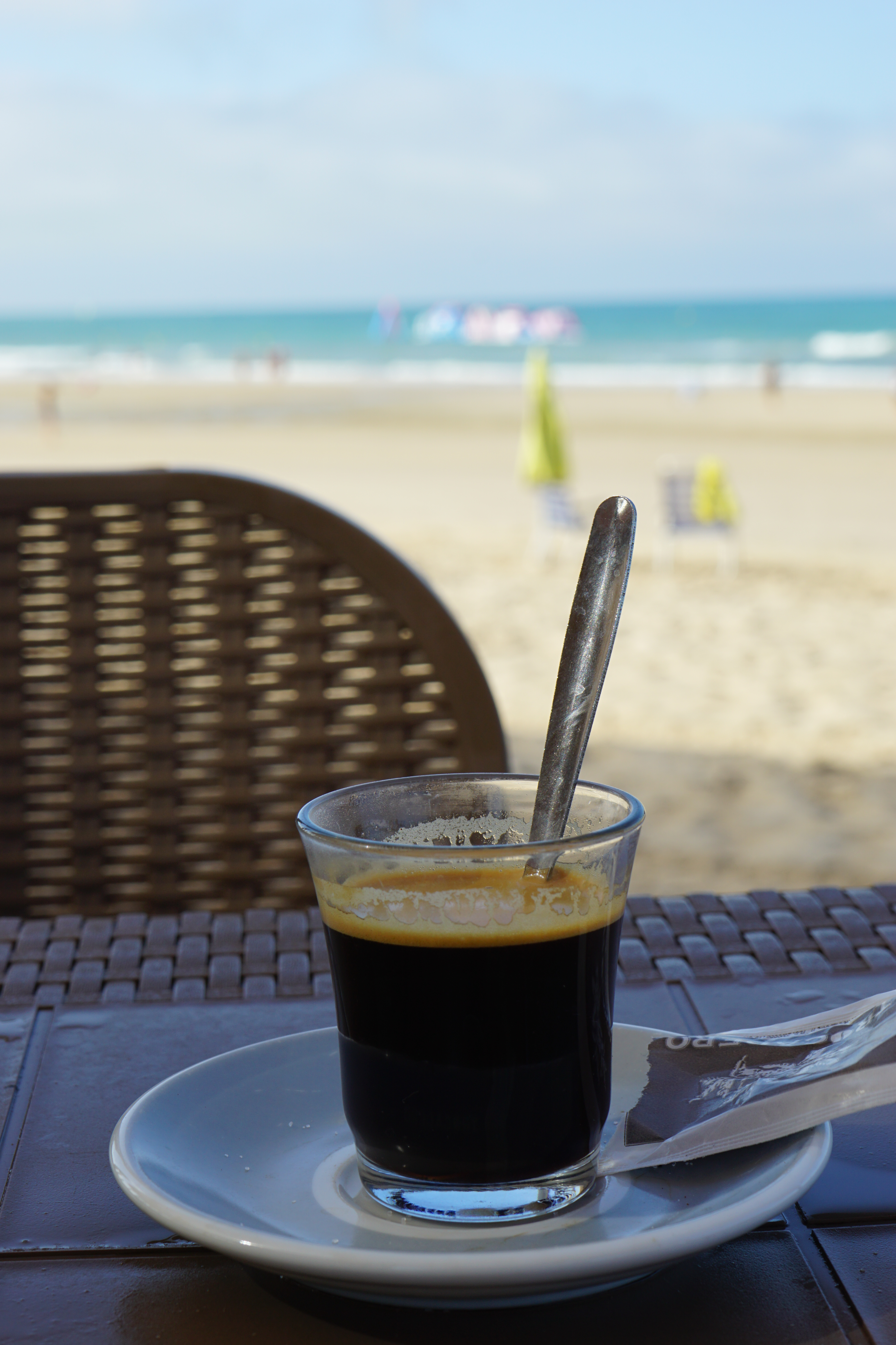 A cup of espresso on a saucer with a teaspoon inside, placed on a wicker table with a blurred background