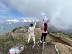 Two people standing on a mountain trail with their backs to the camera, both pointing upward toward the sky. The foreground shows rocky terrain and some scattered belongings. In the background, there are cloud-covered mountains and a vast landscape extending into the distance.