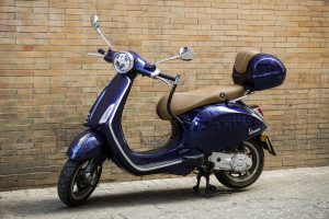 Purple vespa scooter parked in the street, in front of a wall made of bricks