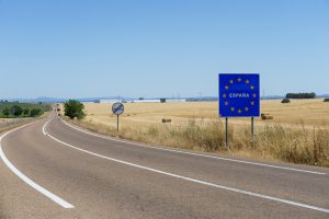 A long, curved road in a rural area with fields on both sides. A blue sign with yellow stars in a circle and the word “ESPANA” (Spain) in the center, indicating the border of Spain
