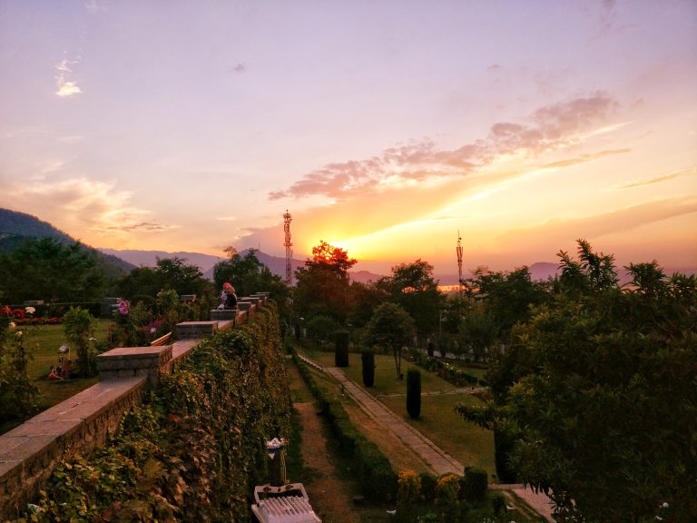 A scenic view of a garden during sunset, with a stone wall covered in greenery on the left. Two people seen sitting on the edge of the wall, looking at the sunset.