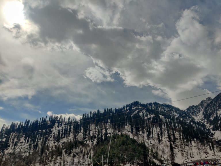 A scenic view of Solang Valley in Manali, India with coniferous trees under a partly cloudy sky, power lines stretching across the foreground.