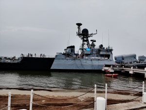 INS Khukri DIU, a large naval ship docked in a harbor with several people standing on its deck and on the dock. The ship has various antennas and equipment on top, and there is a small orange boat floating in the water nearby.