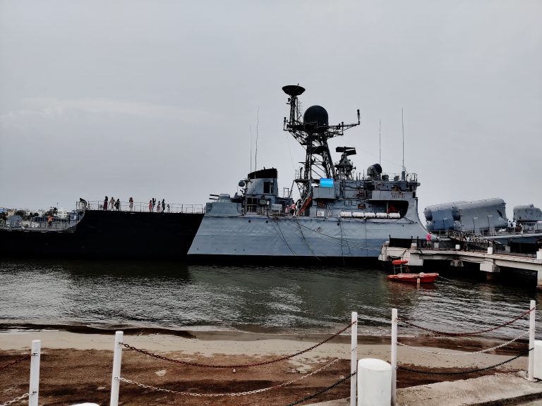 INS Khukri DIU, a large naval ship docked in a harbor with several people standing on its deck and on the dock. The ship has various antennas and equipment on top, and there is a small orange boat floating in the water nearby.