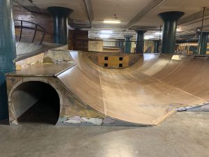 A building interior with a multi-level half-pipe skate ramp with a tunnel under the left side with railings and pillars around the interior space (City Museum, St. Louis, Missouri, USA)
