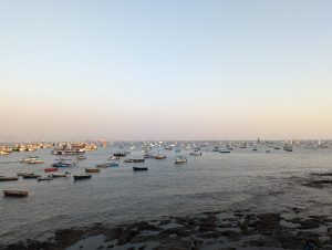 A large number of boats are anchored in a calm sea during early evening, seen from the Gateway of India. The sky is clear, with a subtle gradient from light to darker blue. The foreground features a rocky shore extending into the water of the Arabian Sea 