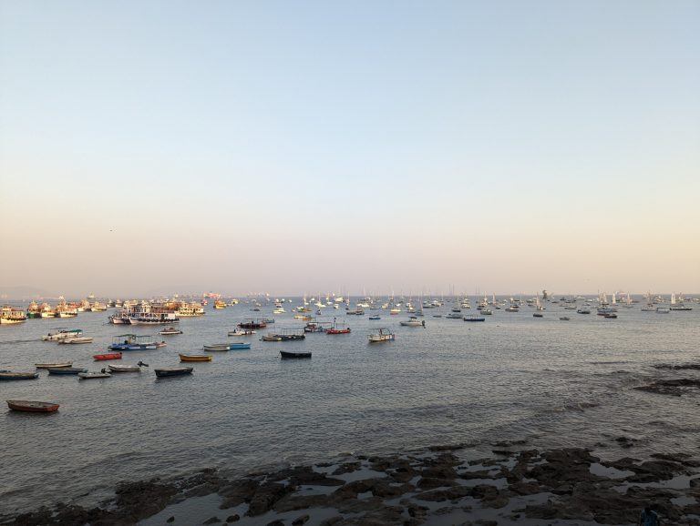 A large number of boats are anchored in a calm sea during early evening, seen from the Gateway of India. The sky is clear, with a subtle gradient from light to darker blue. The foreground features a rocky shore extending into the water of the Arabian Sea