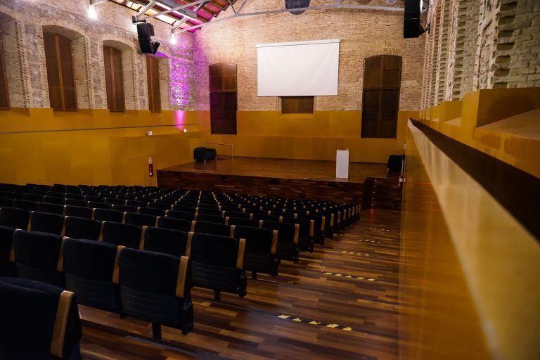 An empty auditorium with rows of black chairs facing a stage. The stage has a white screen, a lectern, and a black chair. The walls are decorated with stone bricks and wooden panels, and there is ambient lighting with a pink hue on the left side.