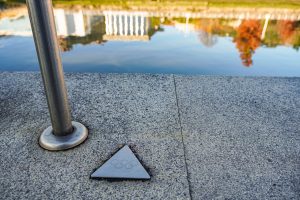 Close-up view of a metal handrail and a triangular plaque with the Olympic rings emblem on a concrete surface near the calm surface of Ria de Bilbao, reflecting buildings and trees on the opposite bank.