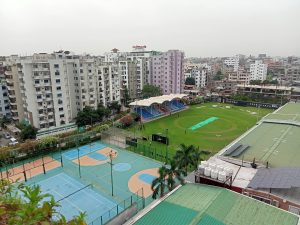 Aerial view of the sports complex at American International University, showing a green cricket field with a small pavilion and covered seating area on one side. There are people visible on the cricket field. Adjacent to the cricket field, there are multiple blue and beige basketball and tennis courts. 