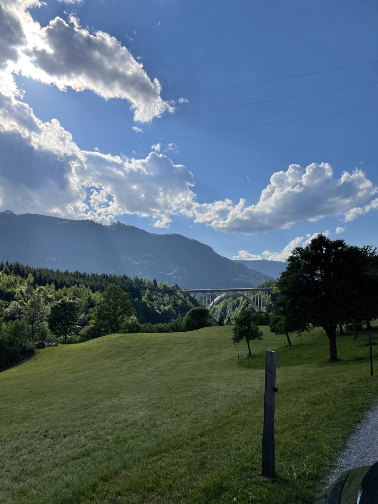 A serene landscape featuring a green field, trees, and rolling hills leading to a distant forest. In the background, there’s a bridge spanning a lush valley with mountainous terrain rising beyond. The sky above is vibrant blue with scattered, fluffy clouds partially obscuring the sun.