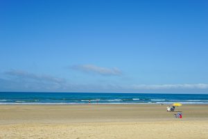 Wide sandy beach with a few people, an umbrella, and beach chairs under a clear blue sky with scattered clouds.