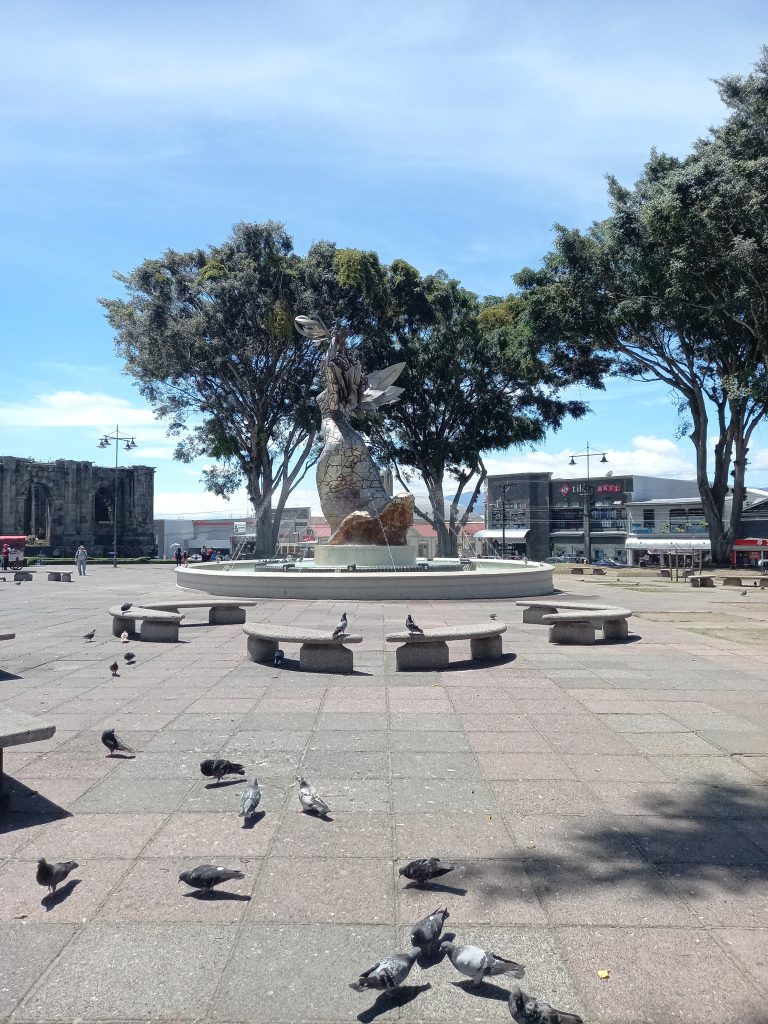 Fountain of the Ruins Park, Cartago, Costa Rica: Urban square with a central monument, trees, pigeons, under a clear blue sky.