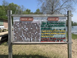 Lakeside wooden sign with Check Out and Check In sections with each having a 20x16 grid with circular tags hanging in various squares (YMCA Camp Eberhart on Corey Lake, Three Rivers, Michigan)