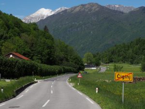 Lush green valley with a small house on the left, dense forest on either side, and snow-capped mountains in the background. A red triangular road sign and a yellow town exit sign labeled "Čiginj" are visible along the roadside.