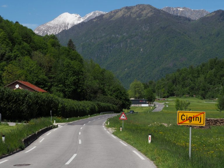 Lush green valley with a small house on the left, dense forest on either side, and snow-capped mountains in the background. A red triangular road sign and a yellow town exit sign labeled “?iginj” are visible along the roadside.