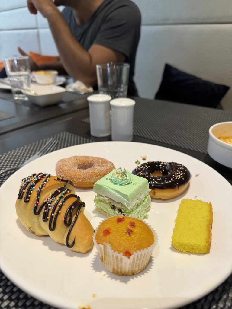 A plate containing various pastries including a croissant drizzled with chocolate and sprinkles, a sugar-coated donut, a piece of green cake with frosting, a donut with chocolate glaze and sprinkles, a yellow sponge cake, and a small muffin with fruit pieces. In the background, a person is seated at the table with glasses, bowls, and a salt and pepper shaker.