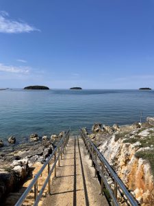  A metal railing pathway leads down rocky steps to a calm, clear sea with small islands in the distance under a bright blue sky.
