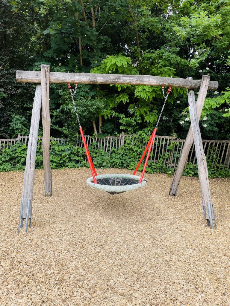 A rustic wooden swing set with red chains and a round, rope net seat is situated in a play area of the Frankfurt Zoo.