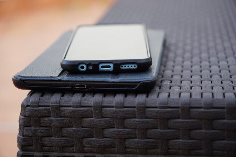 A smartphone and a tablet resting on a woven dark brown outdoor table, with the focus on the charging ports and audio jack.