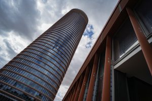 A view from below of a modern, cylindrical high-rise building with a patterned exterior against a dramatic cloudy sky