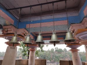 A row of brass bells hanging from the ceiling of a structure with ornate columns. The ceiling is painted in blue and pink stripes