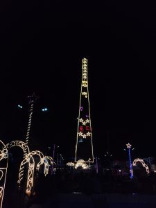 A tall tower-like structure decorated with illuminated star and flower patterns, surrounded by arches also adorned with lights. The scene is set against a dark night sky.