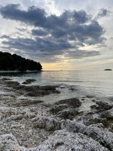  Rocky shoreline with a dramatic cloud-filled sky and calm sea at sunset. Tree-covered landmass visible in the distance on the left.
