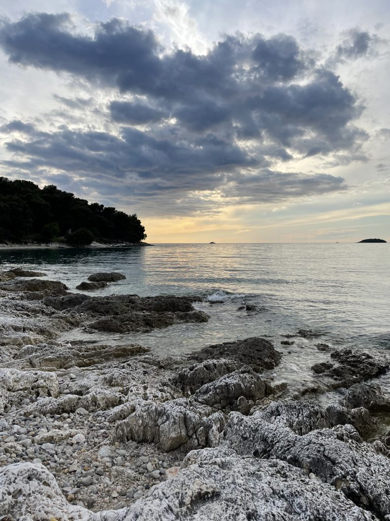 Rocky shoreline with a dramatic cloud-filled sky and calm sea at sunset. Tree-covered landmass visible in the distance on the left.