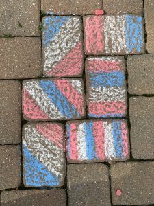 Sidewalk chalk art on a brick pathway featuring a design with diagonal stripes in pink, blue, and white. The bricks also have some moss and grass growing between them.

