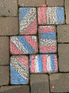 Sidewalk chalk art on a brick pathway featuring a design with diagonal stripes in pink, blue, and white. The bricks also have some moss and grass growing between them.