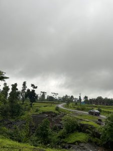 The sky is overcast, with a thick layer of gray clouds. A narrow, winding road cuts through the lush green landscape, leading to a small building in the distance. 