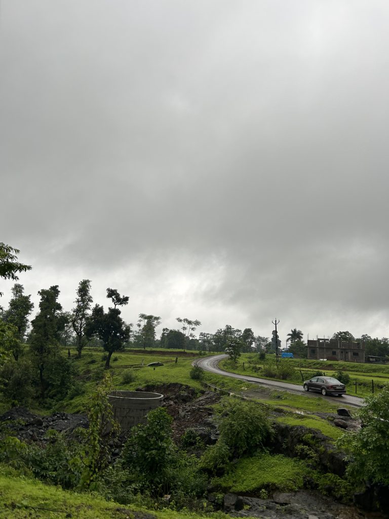 The sky is overcast, with a thick layer of gray clouds. A narrow, winding road cuts through the lush green landscape, leading to a small building in the distance.