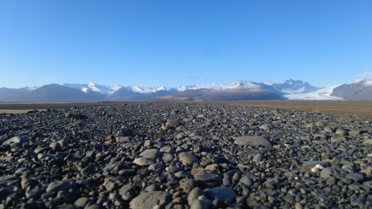 Rocky terrain with scattered stones in the foreground, leading to a flat plain that transitions into towering snow-capped mountains and glaciers under a clear blue sky.