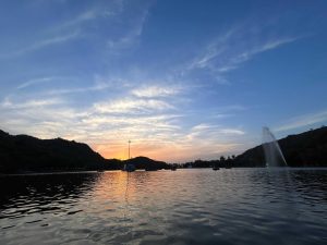 A tranquil lake at sunrise with a fountain spraying water near the center, silhouetted hills in the background, and a colorful sky reflected on the water's surface.