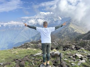 A person stands on a grassy hilltop with arms outstretched, enjoying a scenic view of the majestic mountain range partially covered in clouds under a blue sky on the Great Machhapuchhre Trail at Khumai Dada.