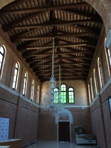 Two chandeliers hanging inside a vaulted two story brick building with wooden truss roof and arched windows (Isola delle Rose, Venice, Italy)
