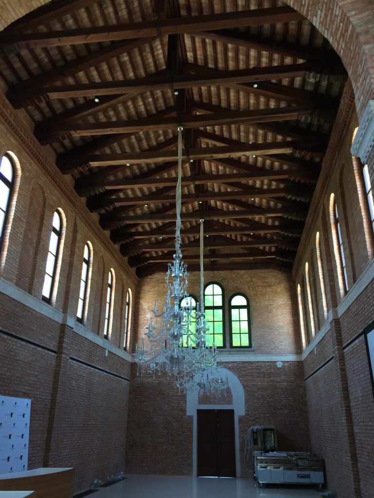 Two chandeliers hanging inside a vaulted two story brick building with wooden truss roof and arched windows (Isola delle Rose, Venice, Italy)