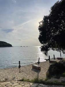 A serene coastal scene with calm sea waters reflecting the sunlight. There are stone pathways leading to the water, lined with benches and closed umbrellas. A large tree is on the right, partially blocking the sun, creating a silhouette effect. In the distance, small boats are floating on the water near the horizon.