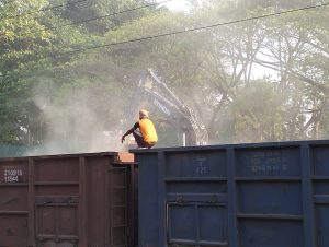 A laborer sitting on a rail rack above a narrow platform, positioned near an excavator's hand, beneath a 25000V DC line during unloading.