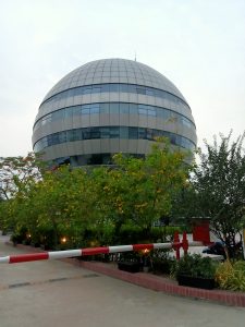 American International University - Bangladesh (AIUB). A spherical building with a checkerboard pattern of glass and opaque panels, surrounded by green trees with yellow flowers. A red and white boom barrier and some planters are visible in the foreground.