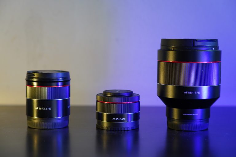 Three camera lenses of varying sizes are displayed on a surface, illuminated by blue and purple accent lighting, with the largest lens on the right. Each lens is marked with its focal length and aperture specifications.