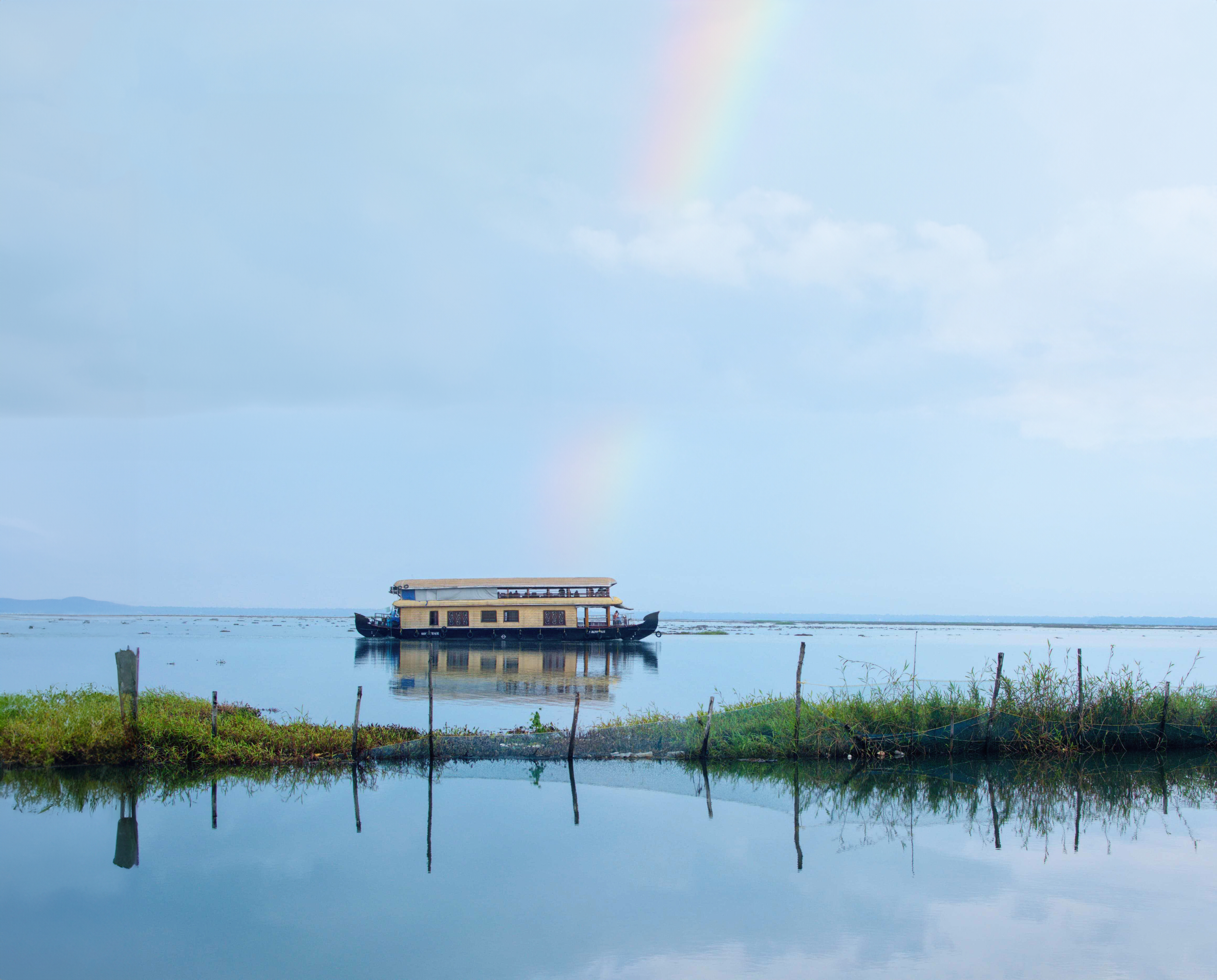 A lakeside view in a monsoon of a houseboat on a calm lake with a faint rainbow in the sky and a small strip of land with grass and wooden stakes in the foreground.