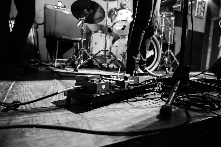 A black and white photo of a live music setup, featuring guitar pedals on the floor, a drum kit, and a musician’s legs and microphone stand in the background.