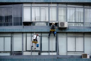 Two workers suspended by ropes in front of a multi-story building. One worker is cleaning windows while the other is servicing an air conditioning unit. Both are wearing safety gear, including helmets and harnesses, and are carrying buckets.